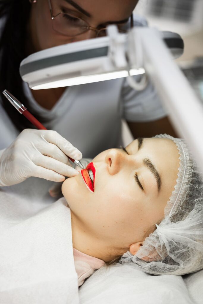 Fachkraft mit Handschuhen tätigt Lippenpigmentierung an liegender Kundin unter Lupenlampe.